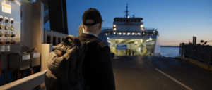 Genevieve Fritschen facing away from the camera and wearing a black hat and backpack walks onto a ferry early in the morning with a sunrise in the background