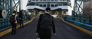 Genevieve Fritschen in a black coat, white shirt and wearing a hat and backpack walks on to the ramp of a ferry