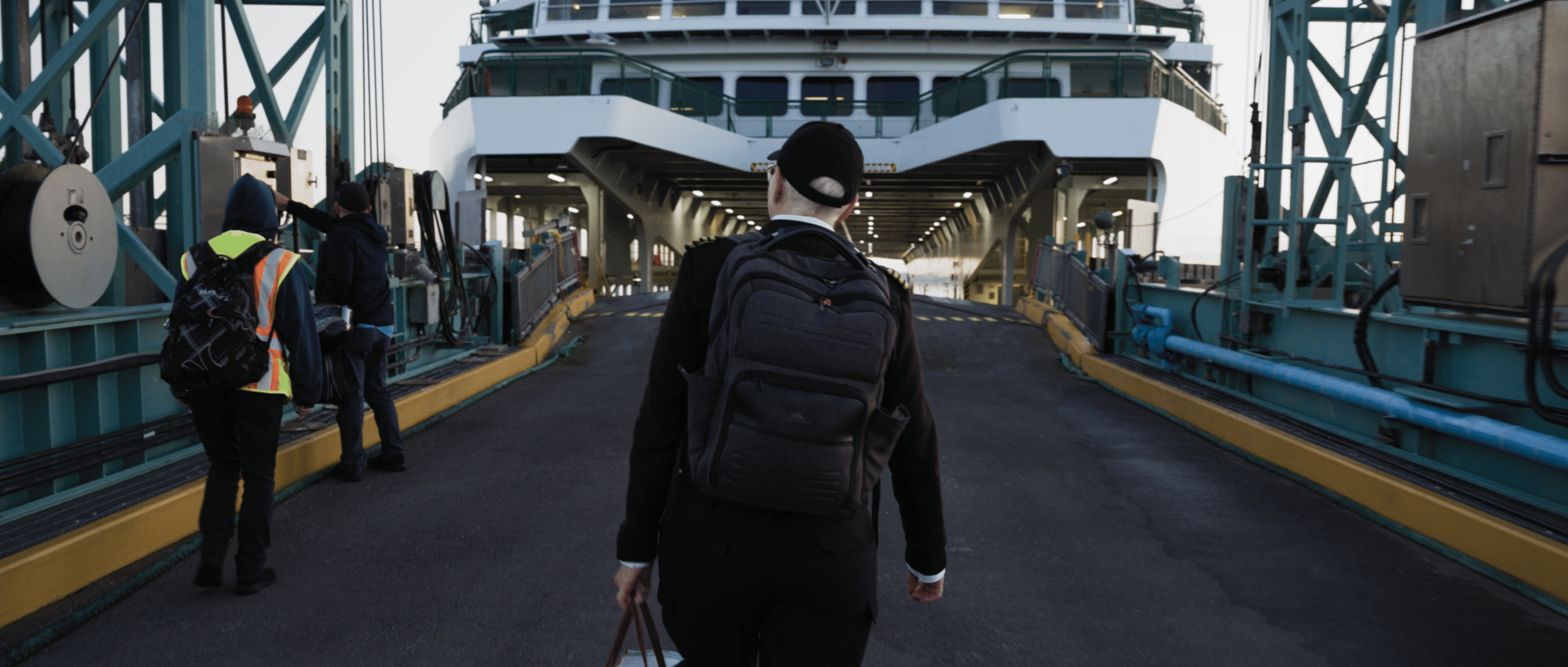 Genevieve Fritschen in a black coat, white shirt and wearing a hat and backpack walks on to the ramp of a ferry