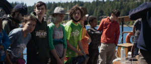 Eight kids look towards the bow of the Adventuress, dressed in shirts and sweaters while the ship is sailing. Green trees and a beach are out of focus in the background.