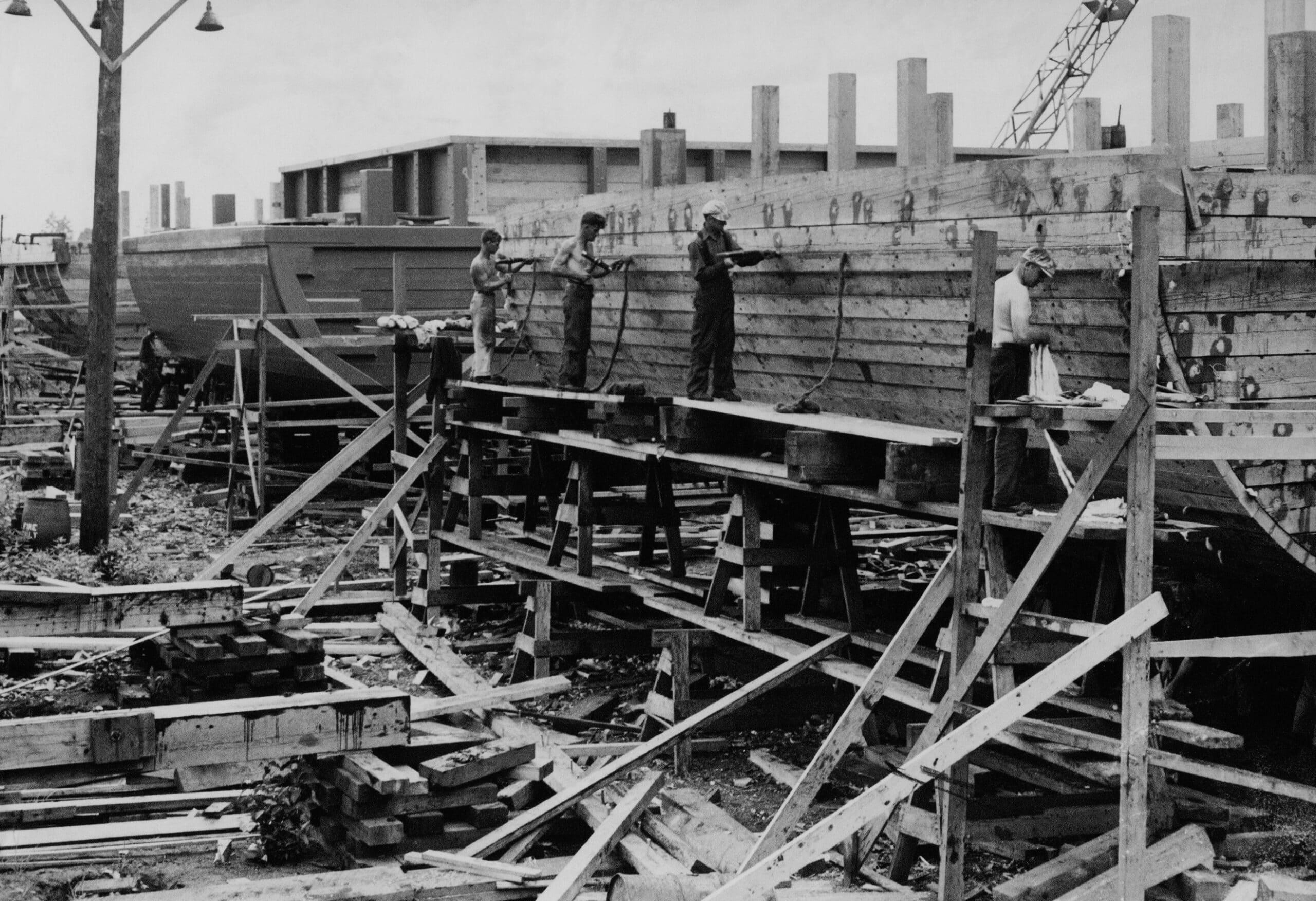 A caulking gang work on scows while standing on scaffolding built from planks and sawhorses. Three scows are partially completed, and the men are hammering the oakum into the nearest boat.