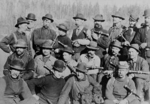 A team of 20 caulkers, all men dressed in a variety of clothes and wearing hats and holding hammers pose for a group photo. Many have thick mustaches.