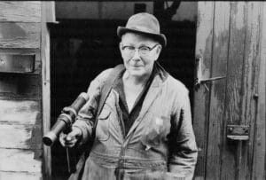 An old man with a hat and overalls holds a specialized caulking hammer while in front of a doorway.