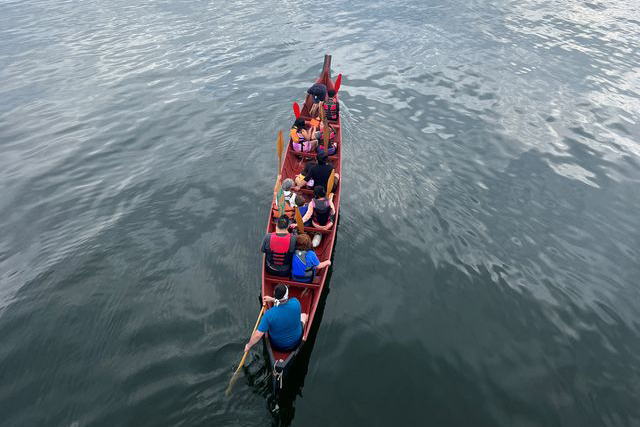An aerial view of a Suquamish canoe gliding through calm waters as pullers raise their paddles into the air.