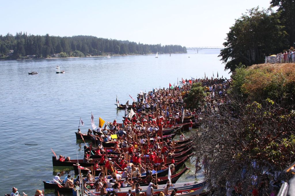 An elevated view of around twenty canoes docked on Suquamish shores, with hundreds of people gathered around them. Agate Pass Bridge is visible in the background.