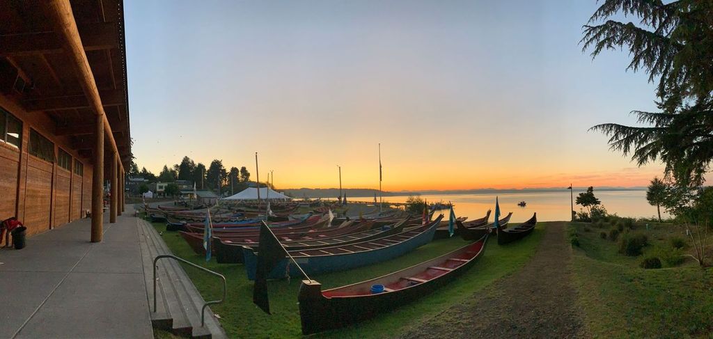 Several canoes laid on the lawn in front of a wooden building, set against sunset over the water.