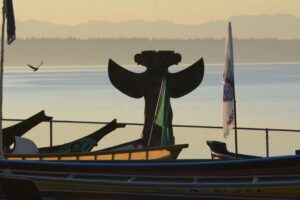 The silhouette of a double-headed eagle memorial pole against the sunset stands behind several docked canoes.