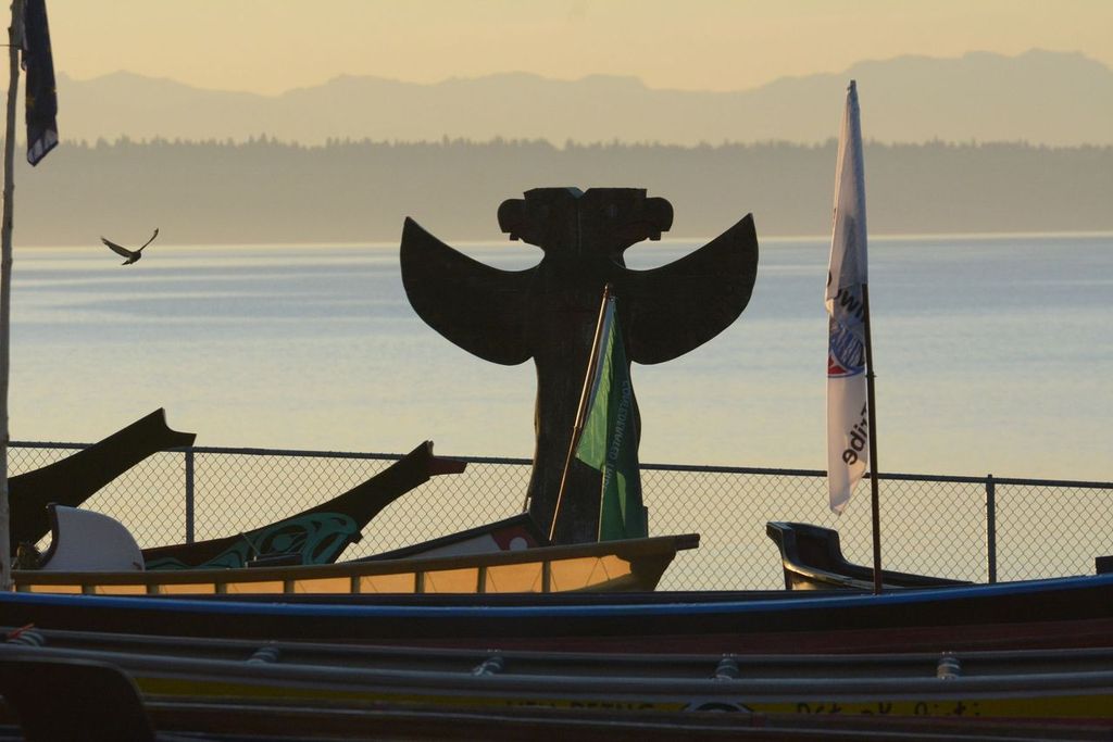 The silhouette of a double-headed eagle memorial pole against the sunset stands behind several docked canoes.