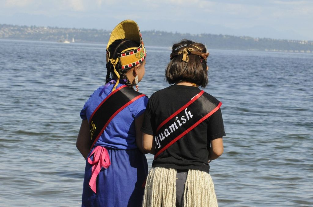 Two children in regalia stand with their backs to the camera, each wearing a sash. One sash reads "Suquamish."