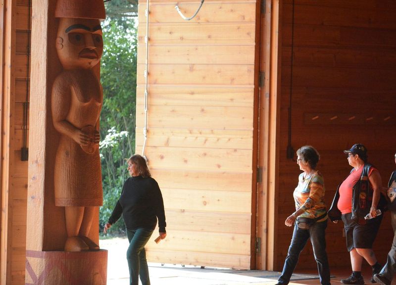 Several people walk in front of a wooden welcome pole within the longhouse-style House of Awakened Culture.