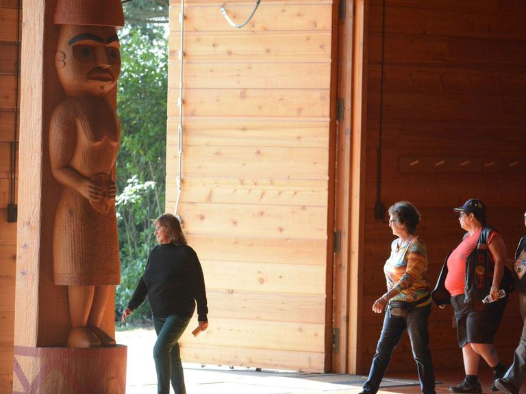 Several people walk in front of a wooden welcome pole within the longhouse-style House of Awakened Culture.