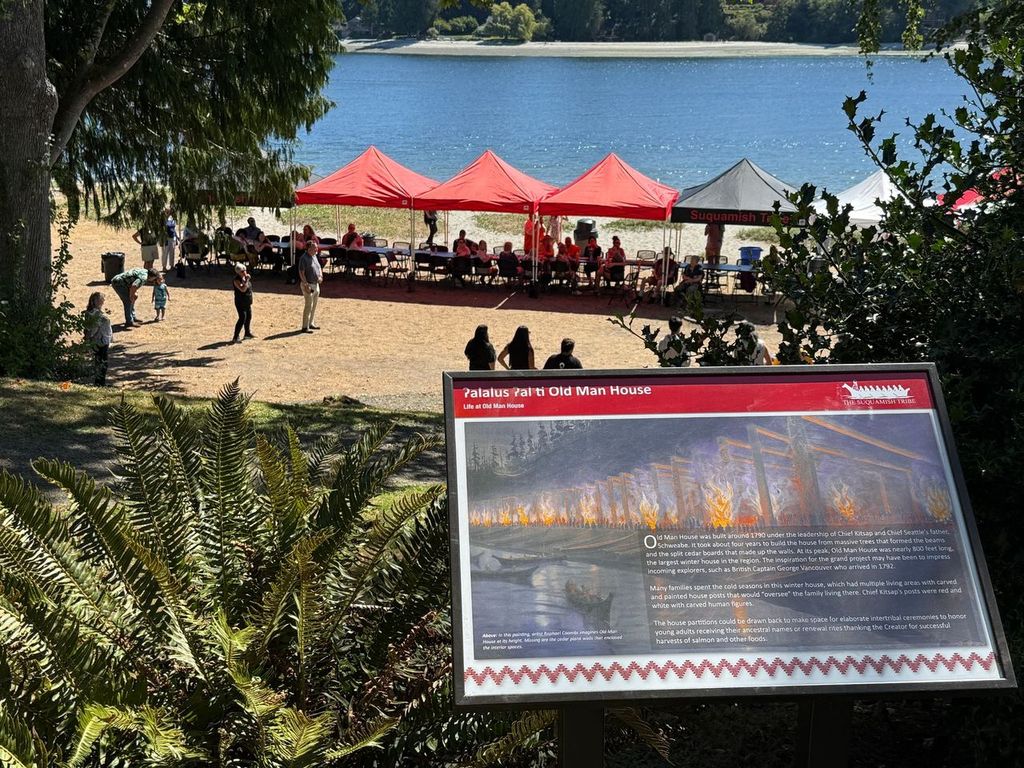 A sign describing the history of Old Man House in the foreground, with canopy tents and people near the shore in the background.