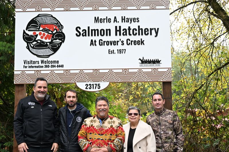 Five adults smile at the camera in front of a large sign reading "Merle A. Hayes Salmon Hatchery at Grovers Creek Est. 1977"