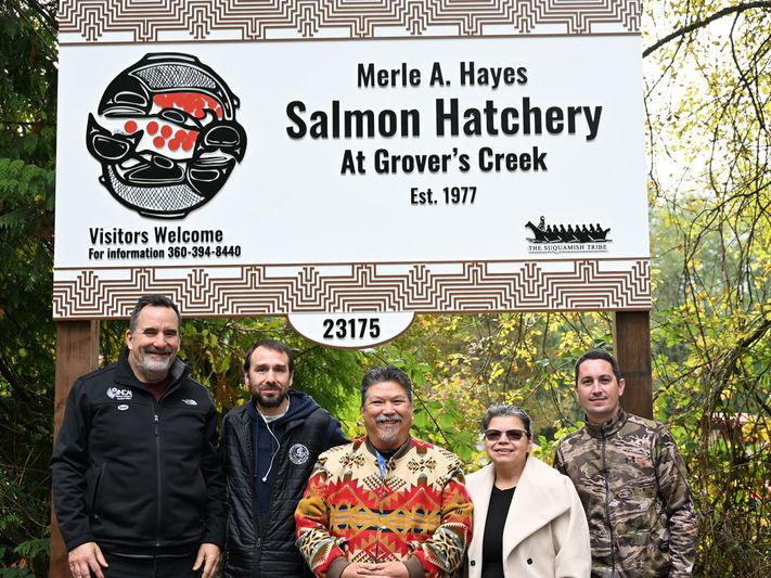 Five adults smile at the camera in front of a large sign reading "Merle A. Hayes Salmon Hatchery at Grovers Creek Est. 1977"