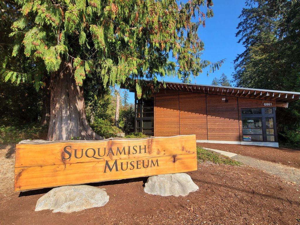 A wooden sign reading "Suquamish Museum" in the foreground, with a longhouse-style museum in the background.