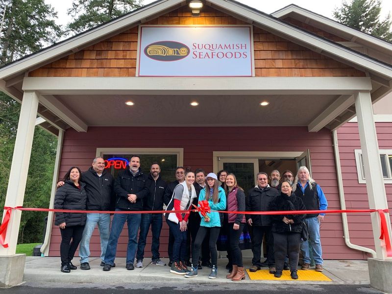 Fifteen men and women stand in front of the Suquamish Seafoods storefront. One woman holds a pair of large scissors as she prepares to cut the red ribbon in front of the group.