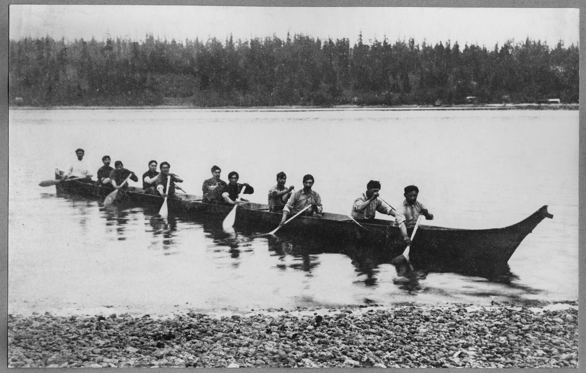 A black-and-white photo of a traditional wooden canoe approaching shore, with eleven men paddling.