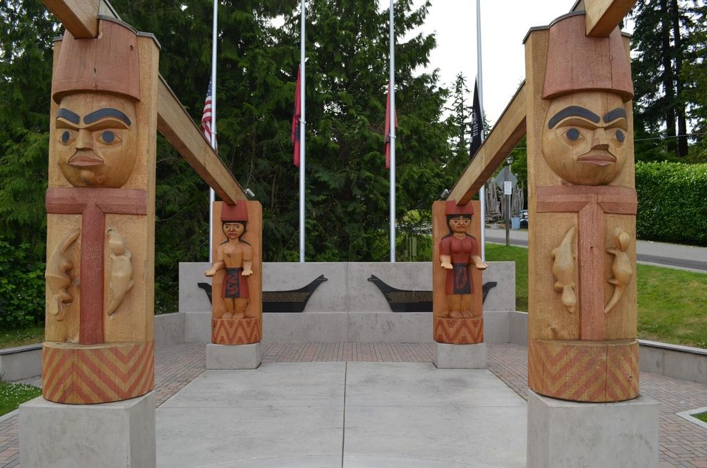 Four carved wooden figures stand on pavement, with two metal canoes decorating a stone memorial behind them. 