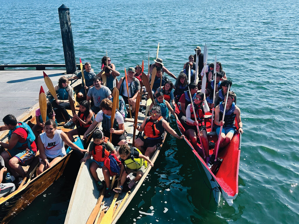 Three canoes gather in the water, full of children and adults wearing colorful life jackets and raising their wooden paddles head-up.