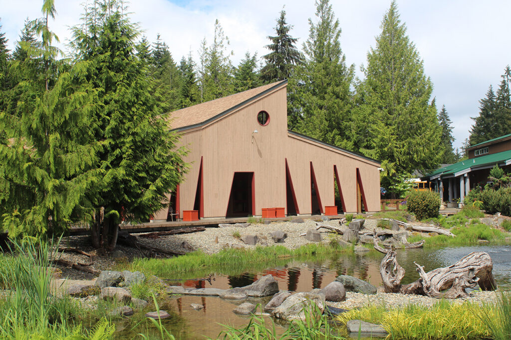 A tan building with sloping roof stands nestled between cedar trees. A small creek is visible in the foreground.