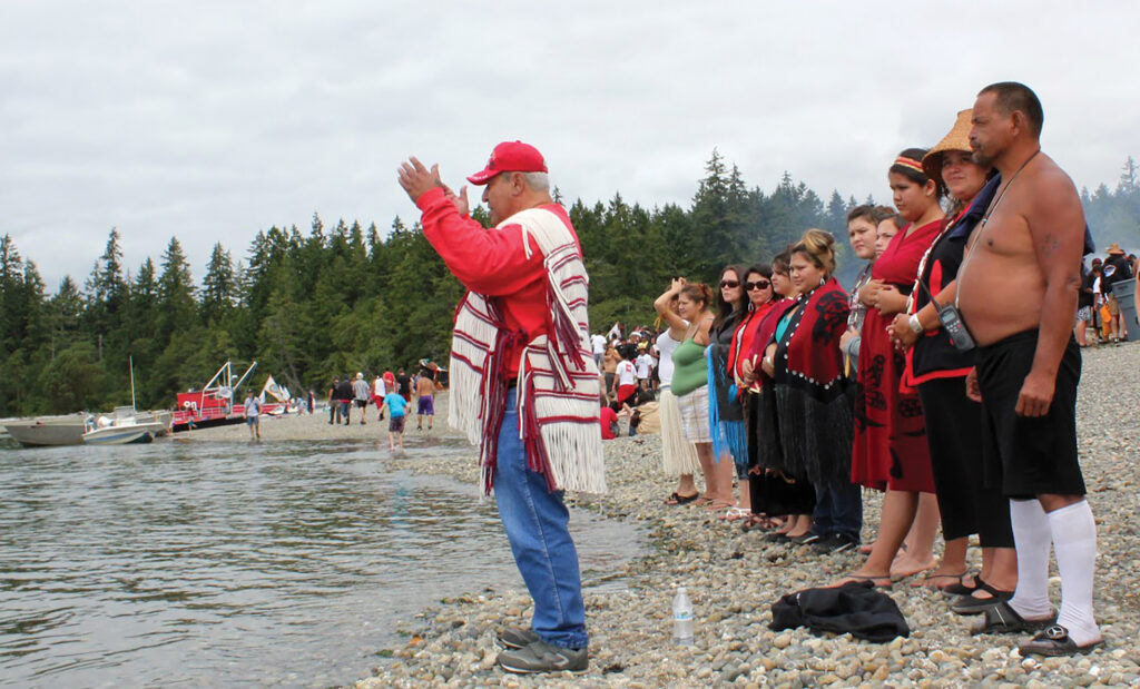 A Squaxin elder man stands facing the shore with hands raised wearing red and white regalia.