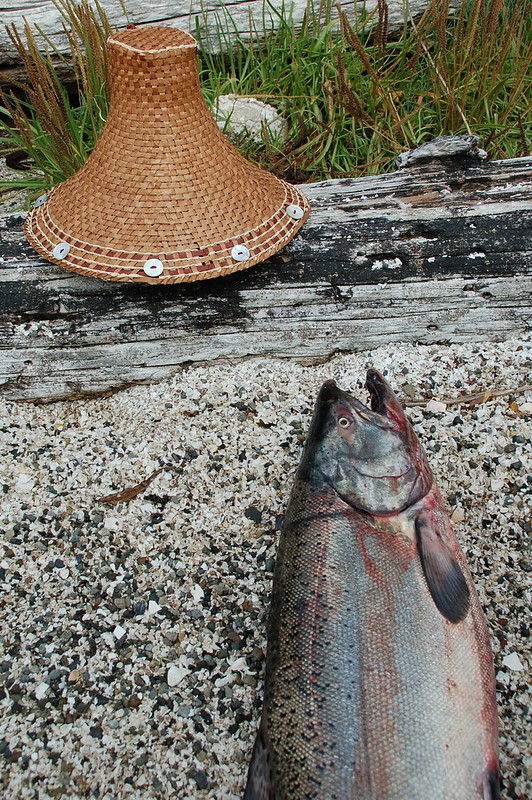 A woven cedar hat with pinto abalone adornments rests on a log in front of a dead salmon.