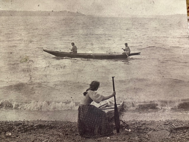 A woman holding a canoe paddle rests on shore, gazing at two men paddling a canoe on the water.