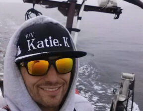 A photo of Duran George smiling at the camera on a boat wearing a hat, hoodie and sunglasses.