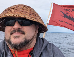 A photo of Nigel Lawrence smiling at the camera, wearing a woven hat and sunglasses, on a boat with the Suquamish flag behind him.
