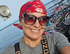 A photo of Shellene George smiling at the camera wearing a bandana and sunglasses taken as a selfie in front of seafood traps.